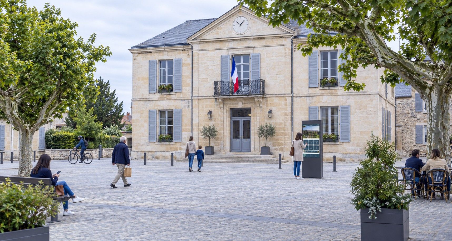 Une place de village animée devant une mairie, où un écran d'information municipale figure parmi les autres éléments de la place